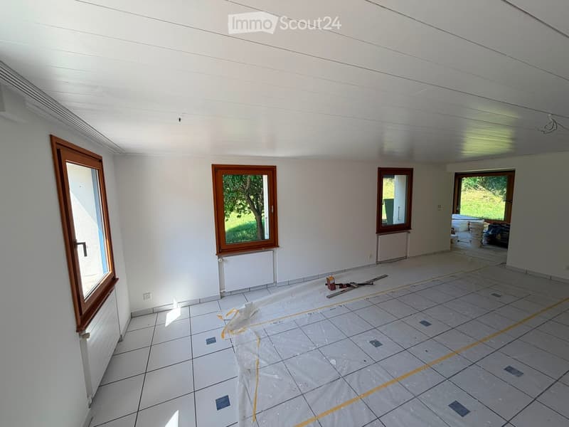Empty room with white walls, tiled floor, and several windows with wooden frames. Some construction materials and tools are visible on the floor.