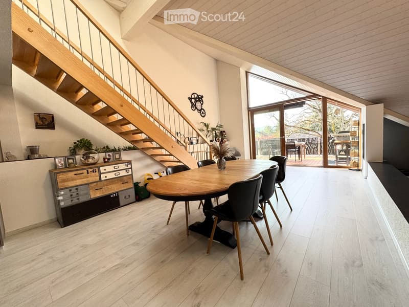 Bright dining area with a round wooden table, six chairs, a wooden cabinet with drawers, and a staircase leading to an upper level.