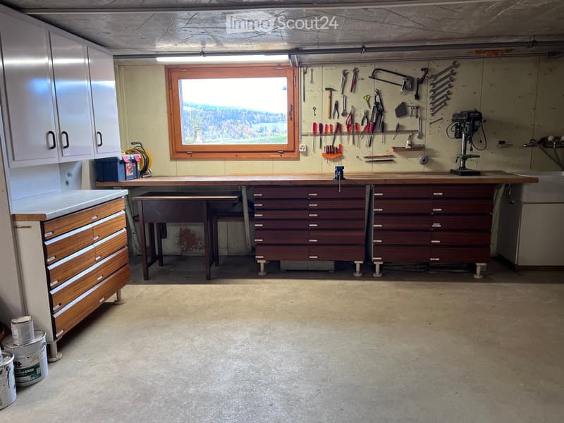 Well-lit workshop with a large window, wooden workbench, tool wall, drill press, and storage cabinets.