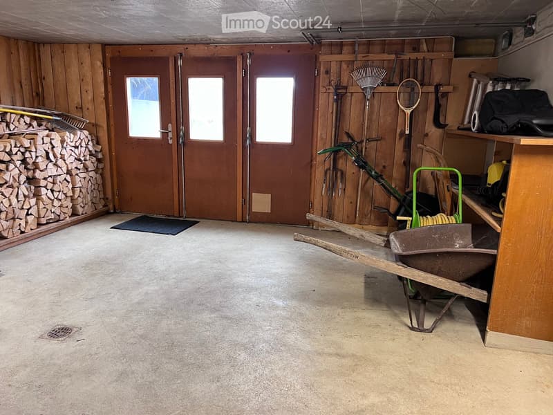 Empty garage with wooden walls, concrete floor, and various tools and equipment stored along the walls, including a wheelbarrow, lawnmower, and gardening tools.