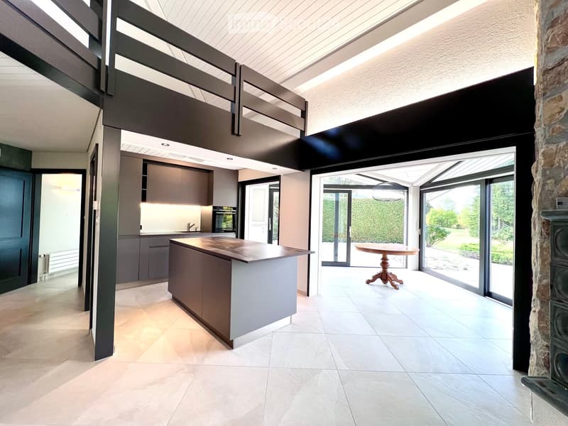 Modern kitchen with grey cabinetry, a central island, and a dining area with a round table and chairs, connected to a spacious living area with large windows and a stone pillar.