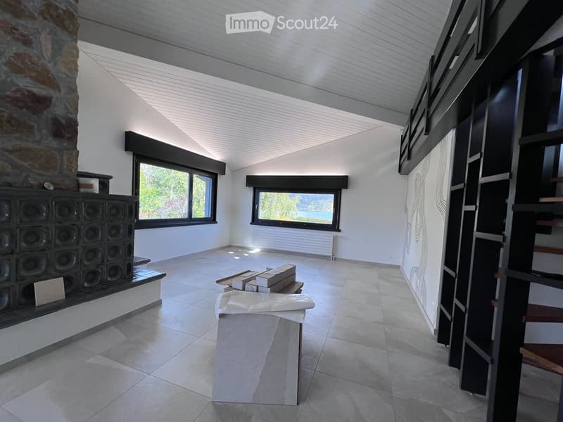 Bright, open room with white walls, tiled floor, large windows, and a stone fireplace. Minimal furnishings include a covered table and stacked boxes.