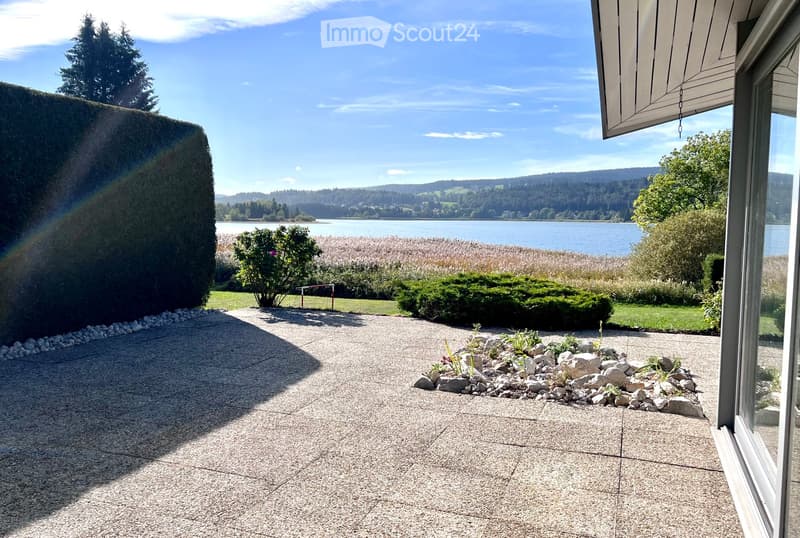 Garden area with gravel pathway, small rock garden, trimmed hedges, and a view of a lake and forested hills in the background.