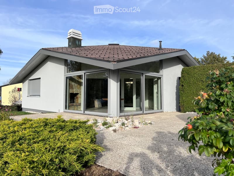 Modern single-story house with white exterior, large glass sliding doors, and a tiled roof. The house features a raised ground floor and is surrounded by neatly trimmed hedges and gravel pathways.