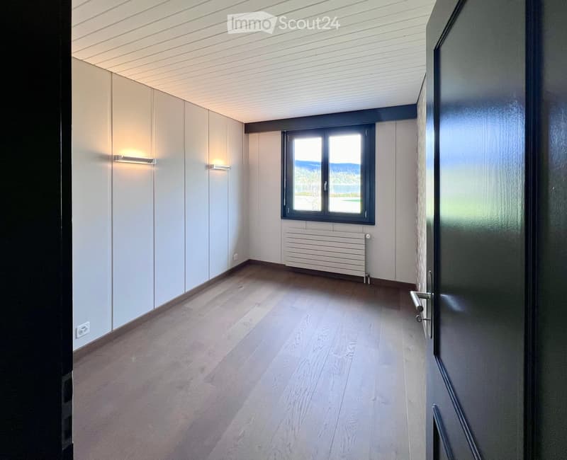 Empty room with light wooden flooring, white walls, a large window with a view, and a radiator. The room has modern lighting fixtures and a closed door.