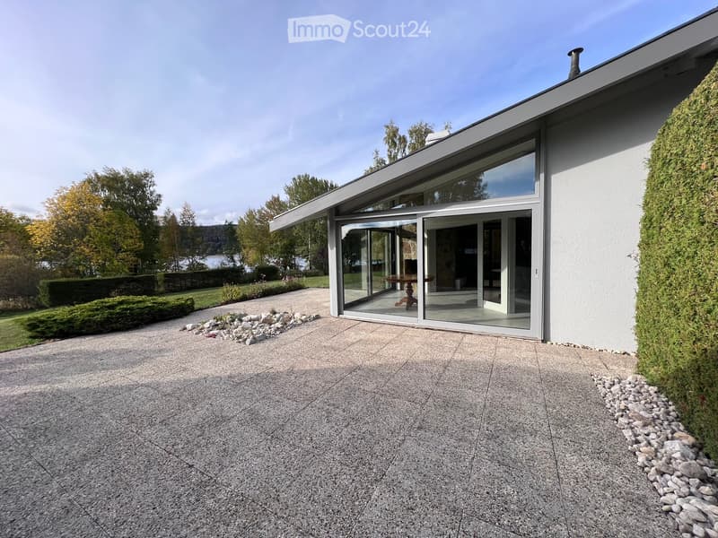 Modern house exterior with large glass sliding doors, minimalist design, and a landscaped garden with a view of a lake.