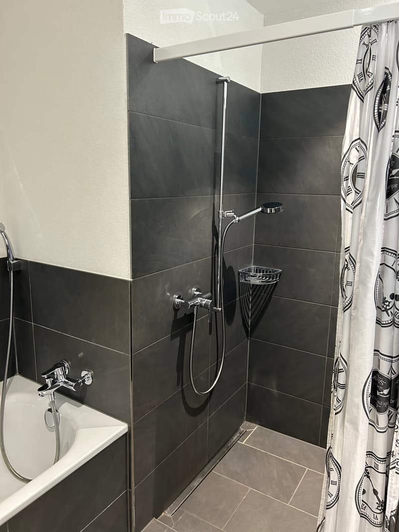 Modern bathroom with dark grey tiles, a bathtub, a showerhead, a shower curtain, and a small shelf for toiletries.
