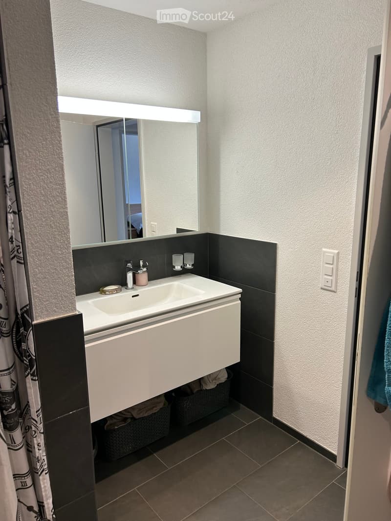 Modern bathroom with a white sink, black tiles, a large mirror with built-in lighting, and a towel hanging on the side.