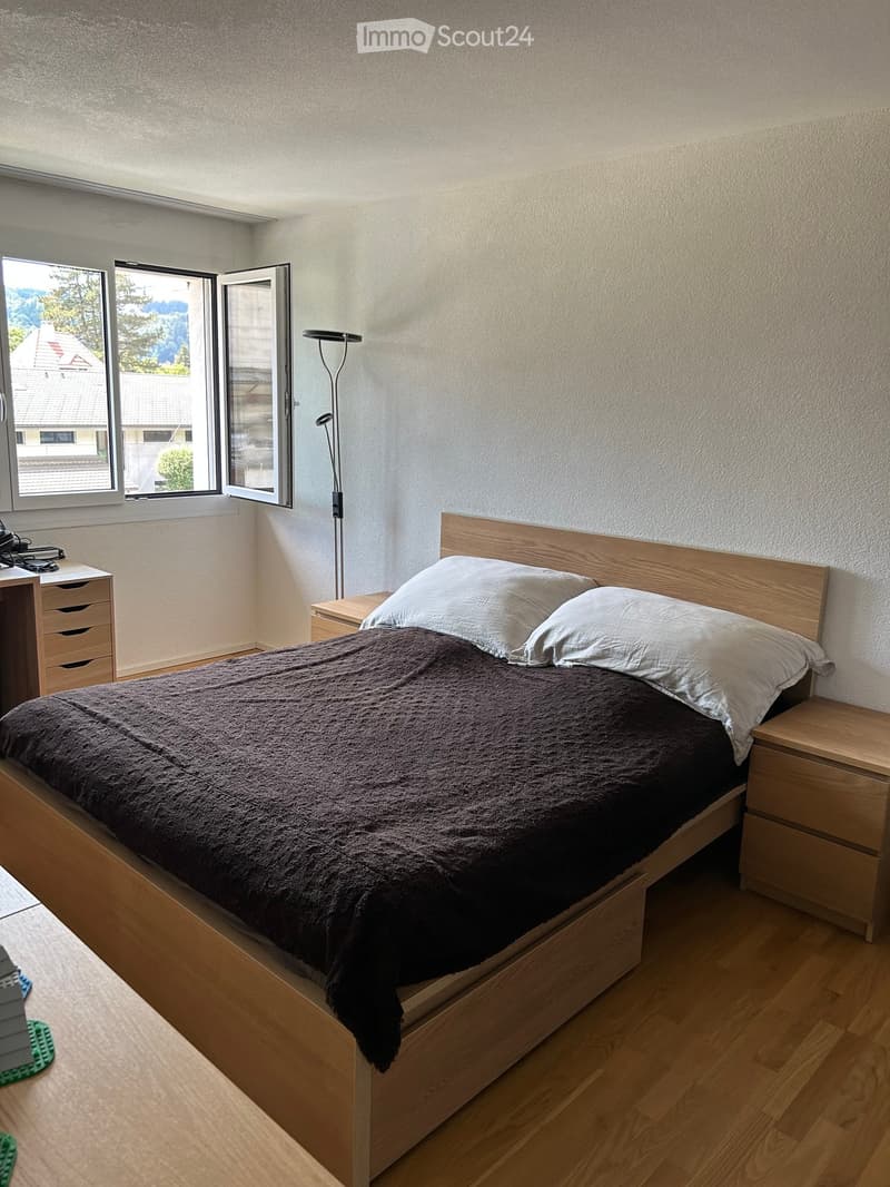 Cozy bedroom with a wooden bed, nightstand, floor lamp, and desk. Large windows offer a view of the surrounding area.