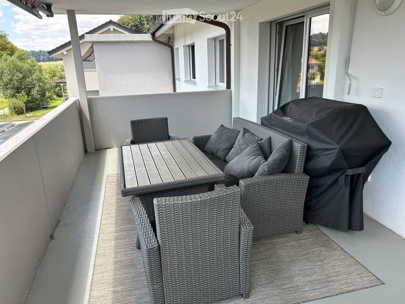 Balcony with wicker furniture, including a sofa, table, chairs, and a covered grill, overlooking a green landscape.