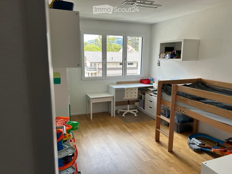 Well-lit bedroom with wooden flooring, bunk bed, desk, chair, shelves, and toys.