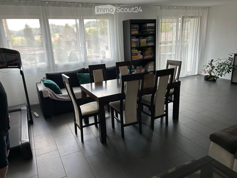 A modern living and dining area with a sofa, dining table, chairs, a bookcase, and an exercise bike. Large windows with sheer curtains let in natural light, and a potted plant adds a touch of greenery.