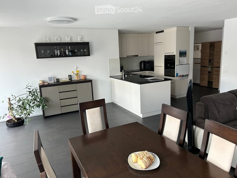 Modern kitchen and dining area with white cabinets, a central island, a dining table with chairs, and a sofa in the background. The kitchen features stainless steel appliances, a black countertop, and a display shelf with bottles and glasses.