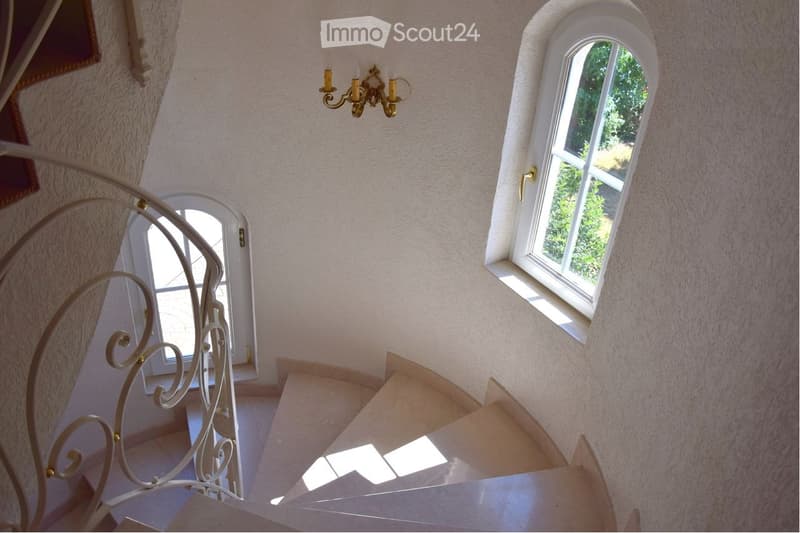 Spiral staircase with ornate metal railing, light fixture with three candles, and arched windows allowing natural light.