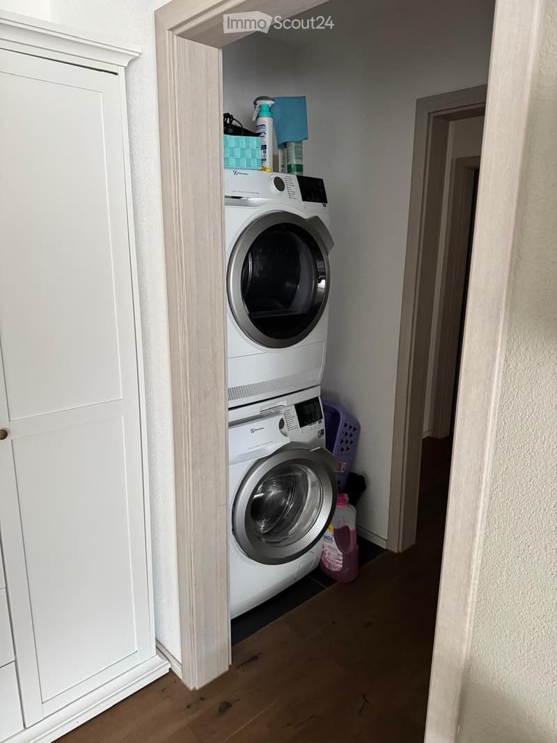 Narrow hallway with a stacked washer and dryer unit, cleaning supplies, and a white cabinet.