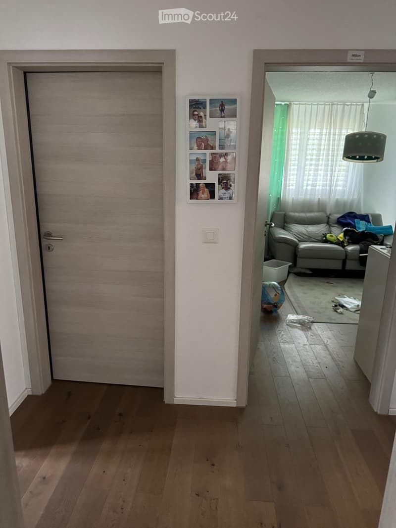 A hallway with wooden flooring, a closed door on the left, and an open doorway leading to a living room on the right. The living room features a grey sofa, a hanging lamp, and a collage of photos on the wall.