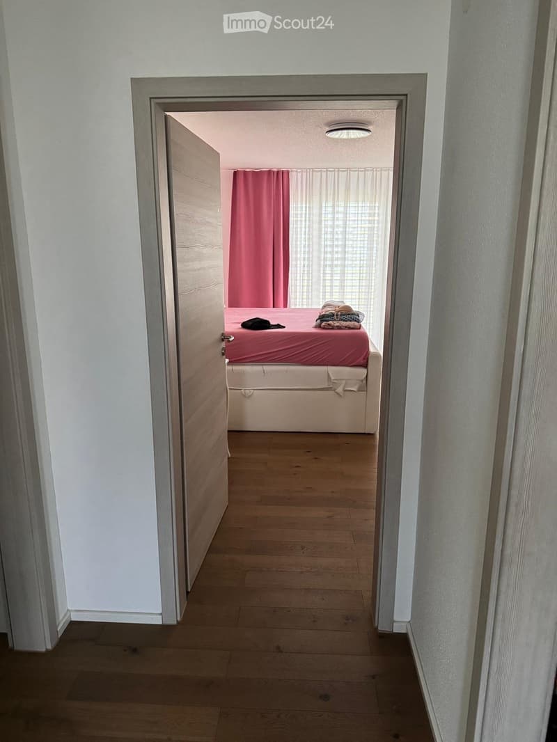 A small bedroom with wooden flooring, a bed with a red bedspread, and a pink curtain on the window.
