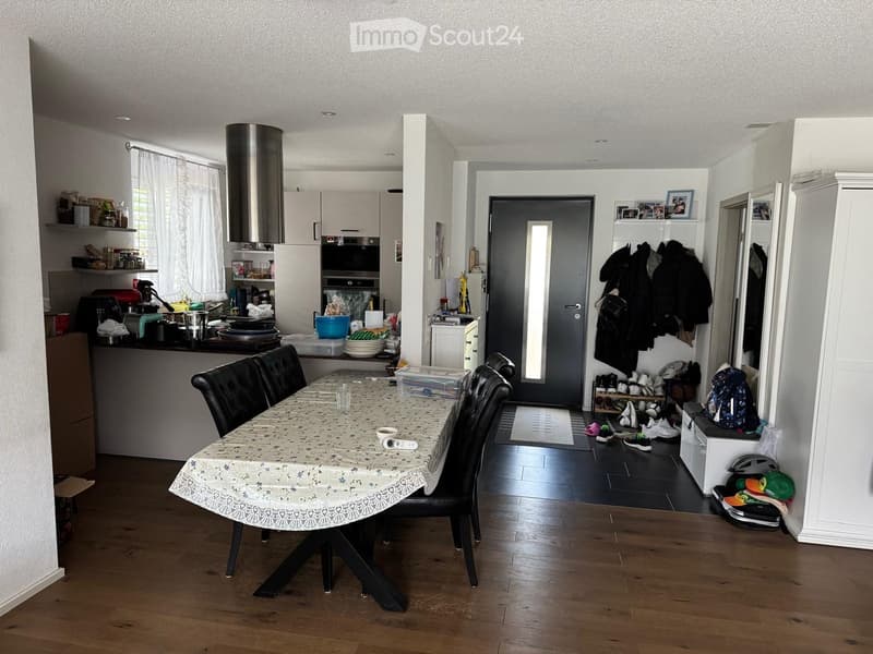 A modern, well-lit room with a dining table and chairs, a kitchen area with appliances, shelves, and a coat rack with jackets and shoes.