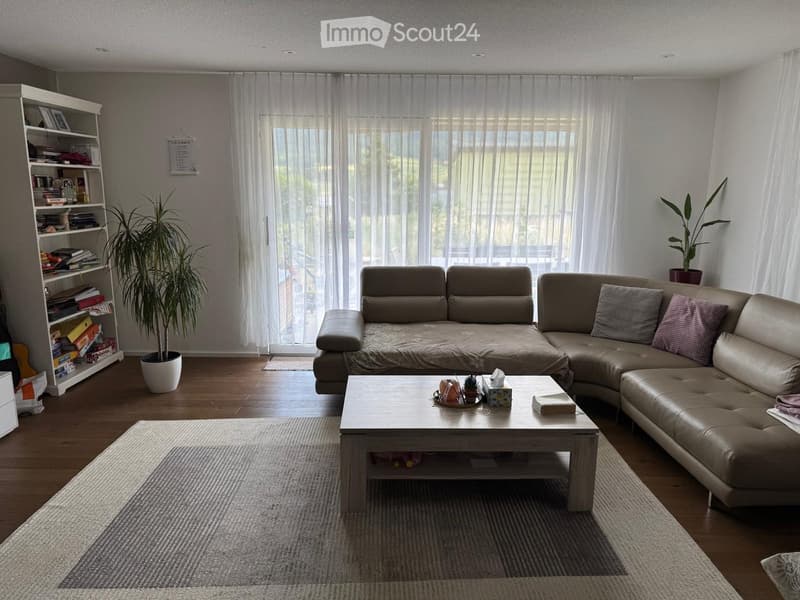 Modern living room with a large sectional sofa, coffee table, bookshelf, and potted plants. The room features wooden flooring and large windows with sheer curtains.