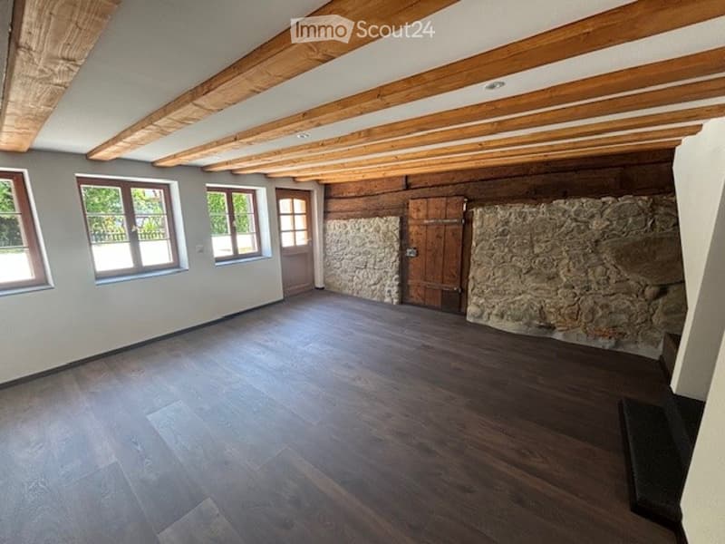 Large open space with exposed wooden beams on the ceiling, stone walls, and hardwood flooring. The room has several large windows providing natural light.