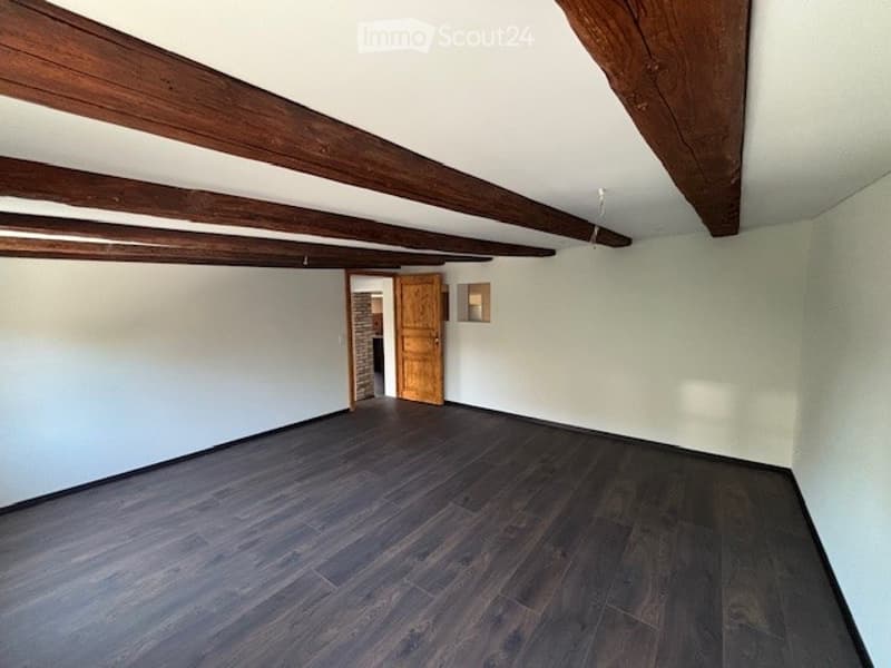 Large open room with exposed wooden beams on the ceiling, hardwood flooring, and a wooden door visible in the background.