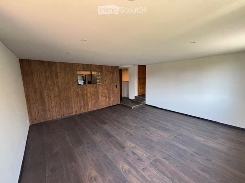 Large open living room with hardwood flooring, a wooden accent wall, and a fireplace visible in the background.