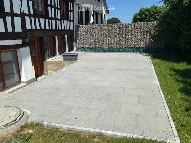 A paved outdoor area adjacent to a traditional half-timbered house, featuring a stone retaining wall and a small planter.