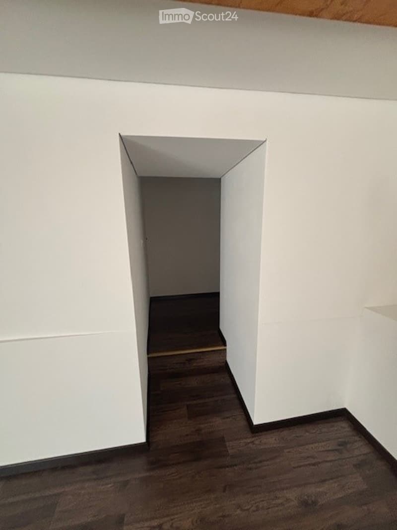 The image shows a room with white walls and a wooden floor. There is a doorway leading to another room or space, with a dark wooden floor visible beyond the doorway.