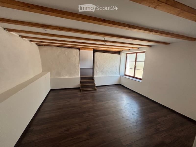 Empty room with wooden floor, white walls, exposed wooden beams on the ceiling, and a small staircase leading to another level.