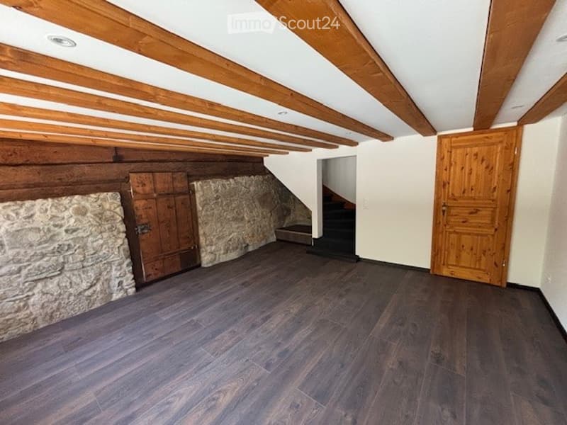 Attic space with exposed wooden beams, stone walls, and a wooden door. The floor is covered with dark hardwood or laminate flooring.