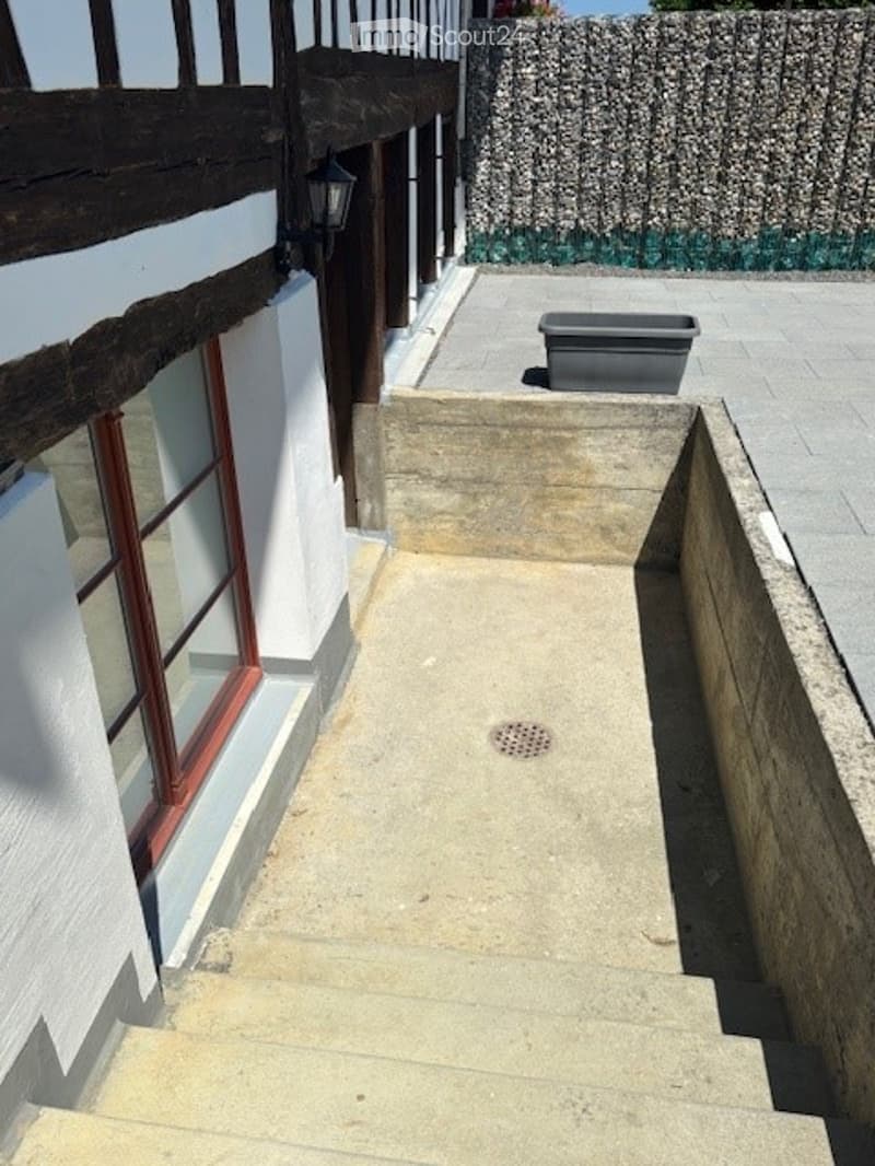 A narrow concrete pathway leading up a set of stairs, bordered by a low stone wall on one side and a building with white walls and red-framed windows on the other. A grey planter sits near the top of the stairs.