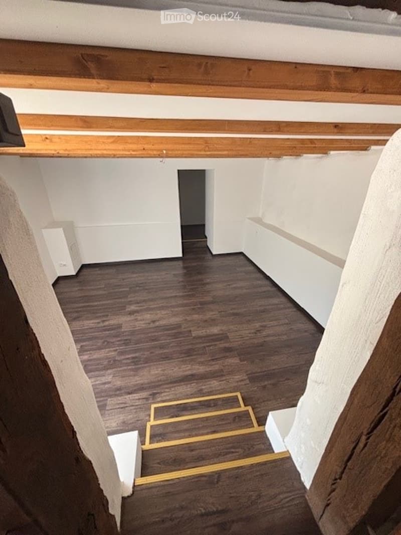 The image shows a room with a wooden ceiling and floor. The room has a doorway leading to another area. The floor appears to be made of dark hardwood or laminate material.