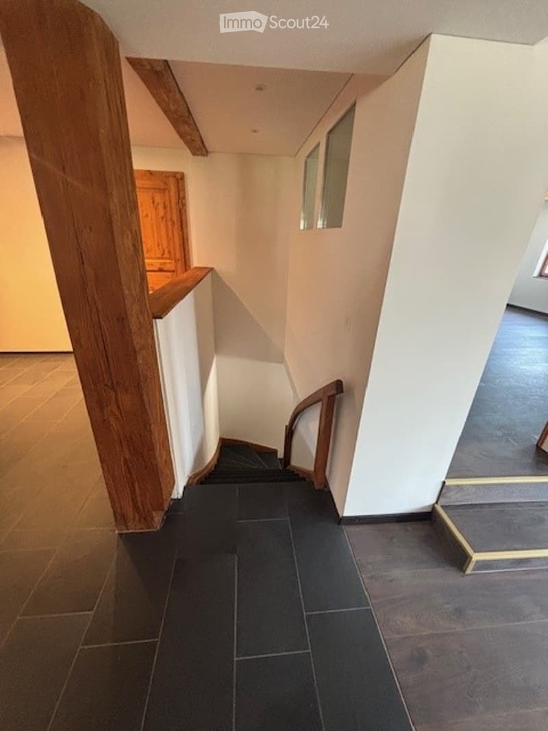 The image shows a hallway with wooden beams, white walls, and a tiled floor. There is a wooden door frame visible, and the hallway leads to another room or area.