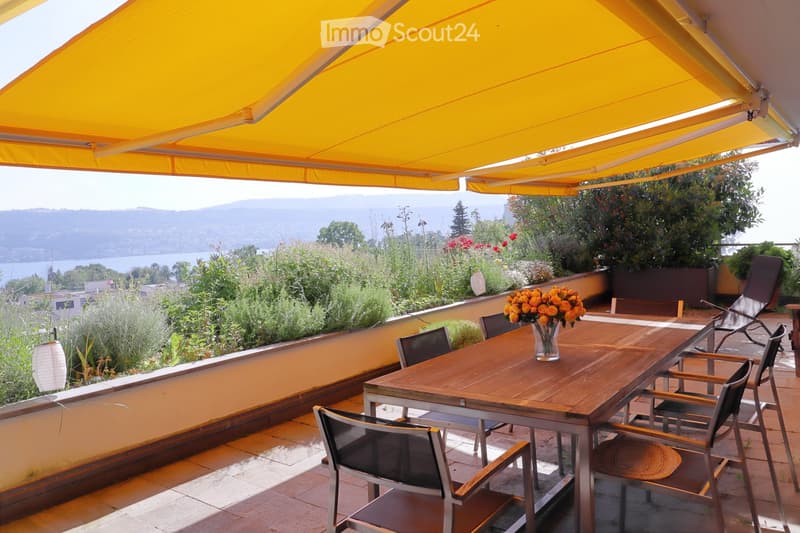 Balcony with a wooden table, chairs, and a vase of orange flowers, overlooking a scenic lake and mountains.