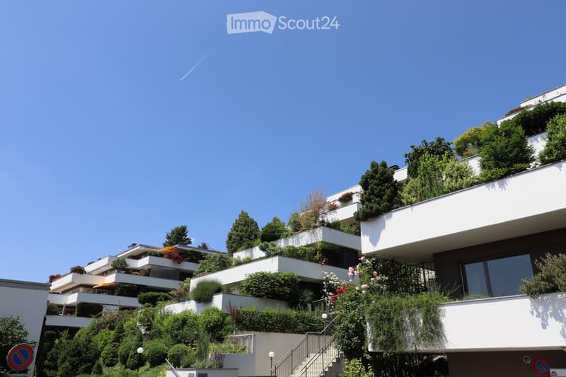 Modern multi-story residential building with terraced balconies and lush greenery, featuring white and dark-colored facades.