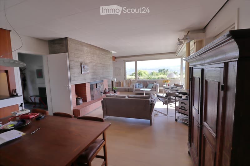 A modern living room with a fireplace, large windows offering a scenic view, a sofa, coffee table, chairs, and a dining table with chairs.