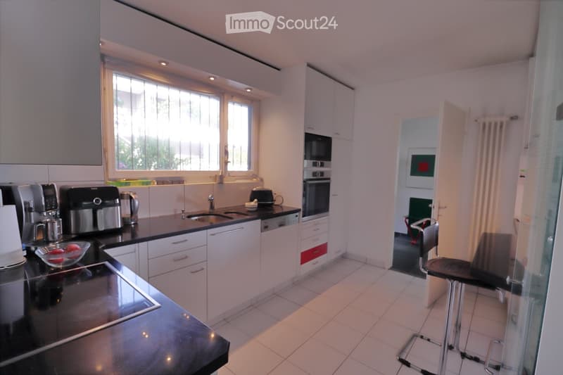 Modern kitchen with white cabinets, black countertops, and stainless steel appliances. Features include a coffee maker, fridge, oven, dishwasher, sink, and a small seating area with a stool and chair.