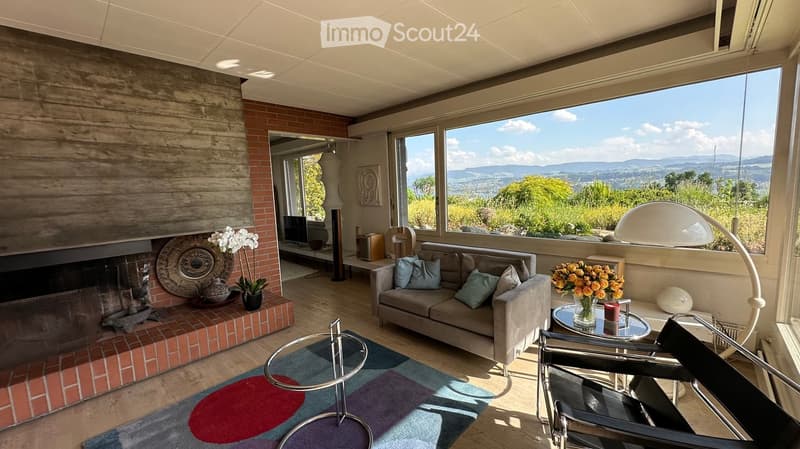 Modern living room with large windows offering a scenic view, featuring a grey sofa, coffee table, fireplace, and decorative vases.