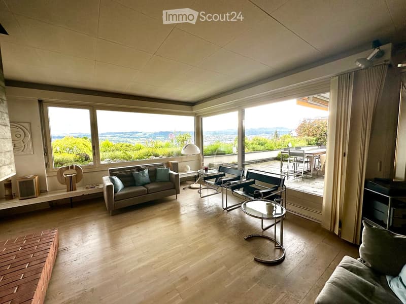 Modern living room with large windows offering a scenic view, featuring a grey sofa, coffee table, chairs, and a lamp.