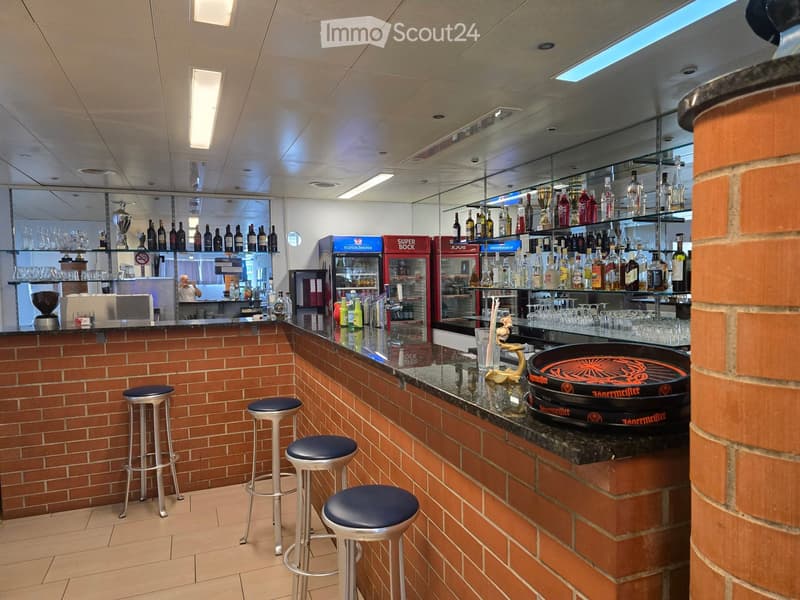 Bar area with brick walls, a counter, four stools, shelves with bottles, and a refrigerator.