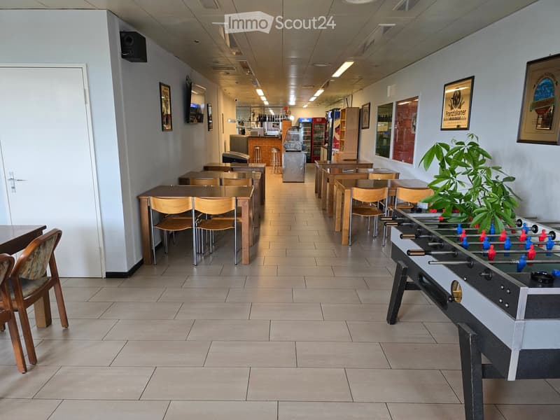 A spacious, well-lit room with tiled flooring and white walls. It features multiple tables and chairs arranged for seating, a foosball table, a small bar area with a refrigerator, and a potted plant near the foosball table.