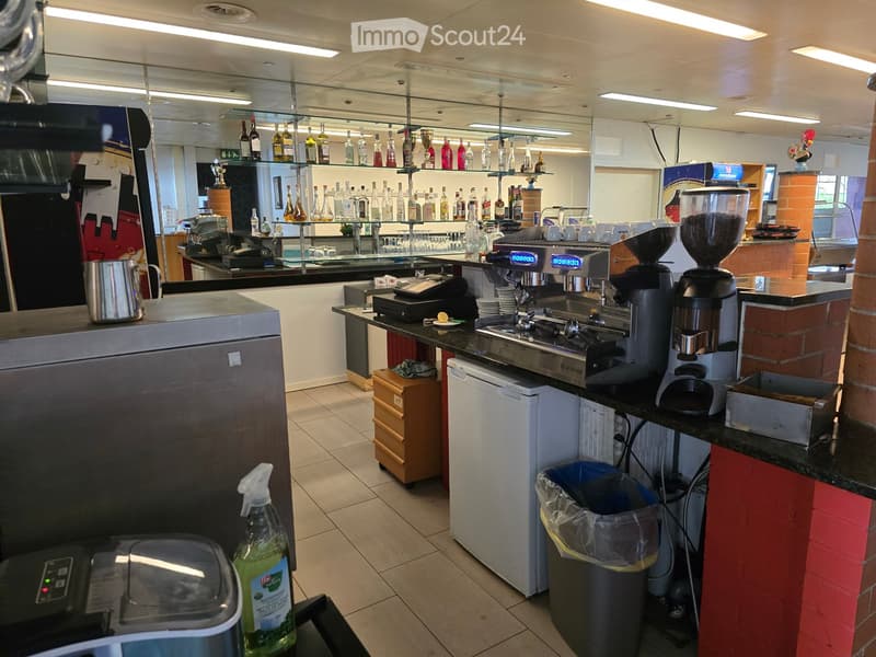 A modern kitchen with stainless steel appliances, including a refrigerator, coffee machine, and dishwasher. Shelves hold various bottles and cups, and a countertop features a sink and additional kitchen equipment.