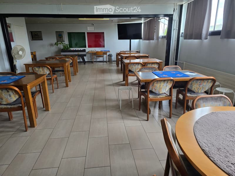 A spacious room with tiled flooring, multiple wooden tables, and chairs arranged in rows. A fan is positioned near the wall, and a large screen is mounted on the wall.