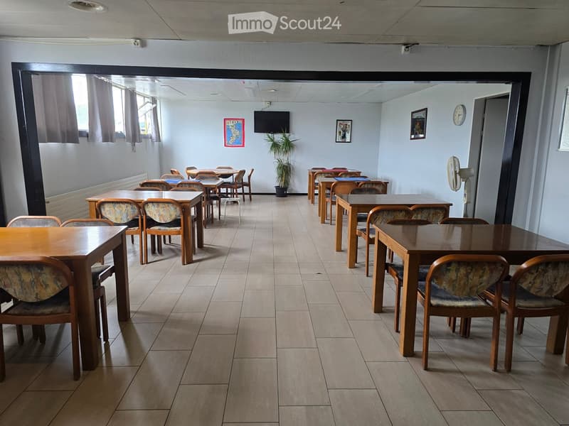 A spacious room with tiled flooring, multiple wooden tables and chairs arranged in rows, a potted plant, a fan, and a television screen mounted on the wall.