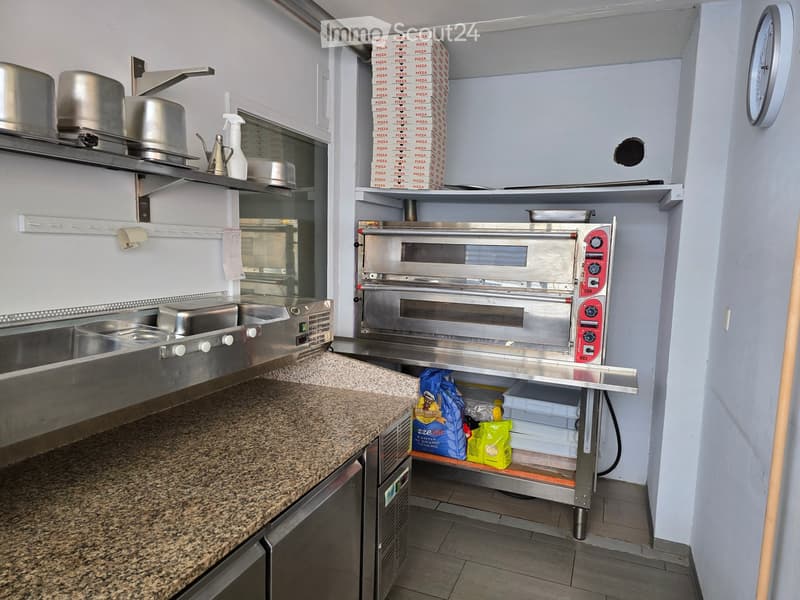 Commercial kitchen with stainless steel oven, granite countertop, shelves holding pizza boxes, and various kitchen equipment.