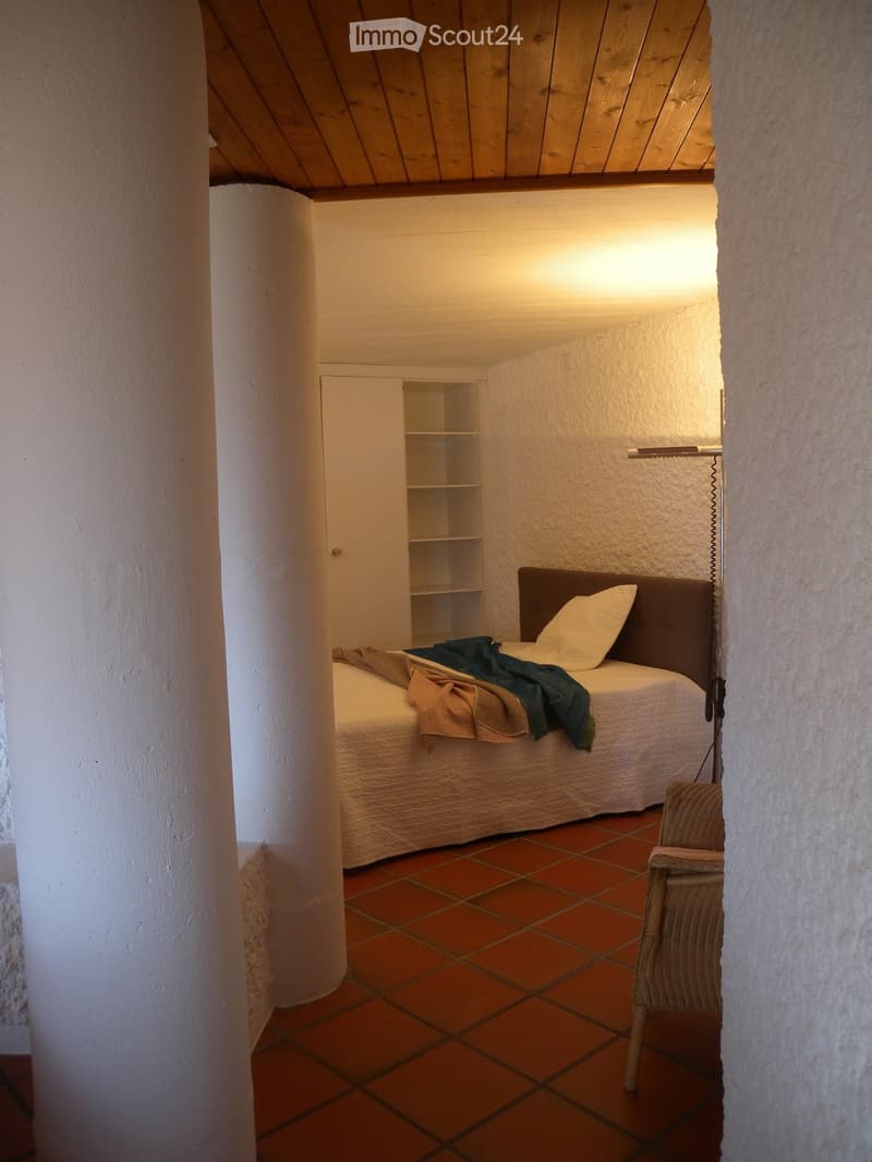 The image shows a cozy bedroom with a wooden ceiling, white walls, and a tiled floor. The room features a bed with bedding, a bookshelf, and a chair.