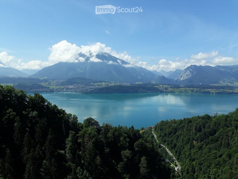 Scenic view of a large lake surrounded by mountains and forests. The lake has a turquoise blue color and is reflected in the calm waters. In the background, there are snow-capped peaks with clouds around them. The landscape is lush and green, with dense f