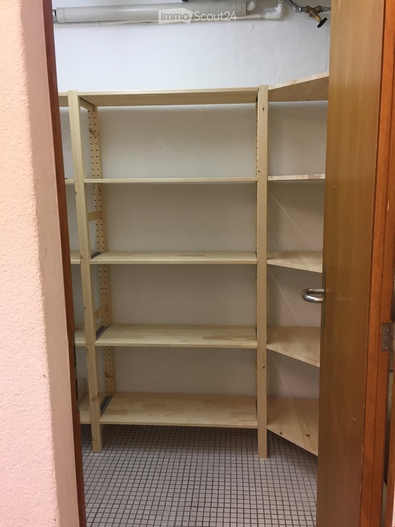 The image shows a storage closet with multiple wooden shelves. The shelves appear to be adjustable and can be used to store various items. The closet has a tiled floor and a wooden door frame.