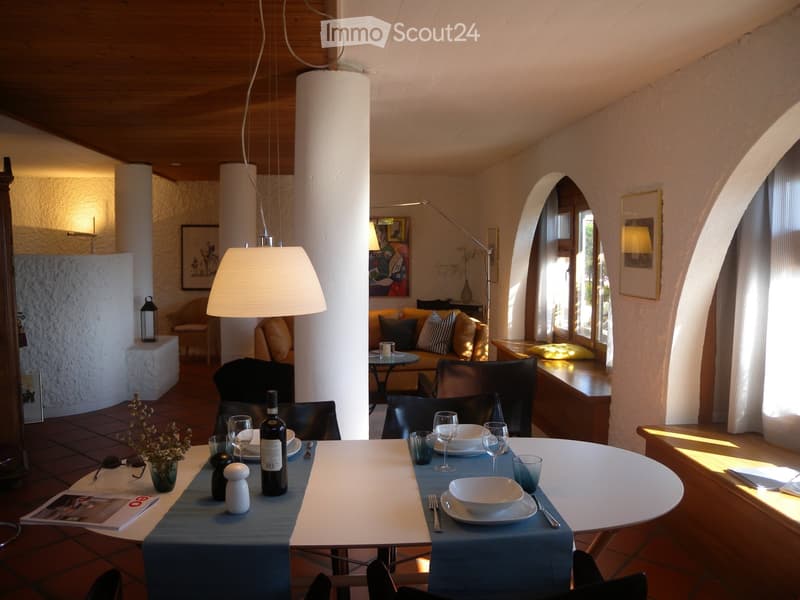 Cozy living room with wooden ceiling, arched doorway, dining table set with plates and wine glasses, and a fireplace visible in the background.