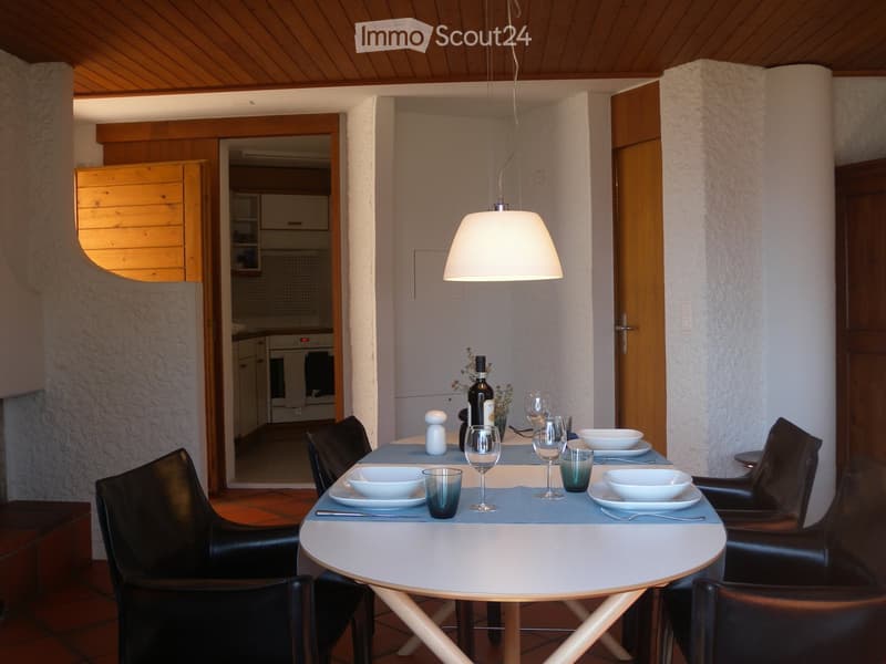 Dining room with a round white table, black leather chairs, a hanging light fixture, and a kitchen visible in the background.